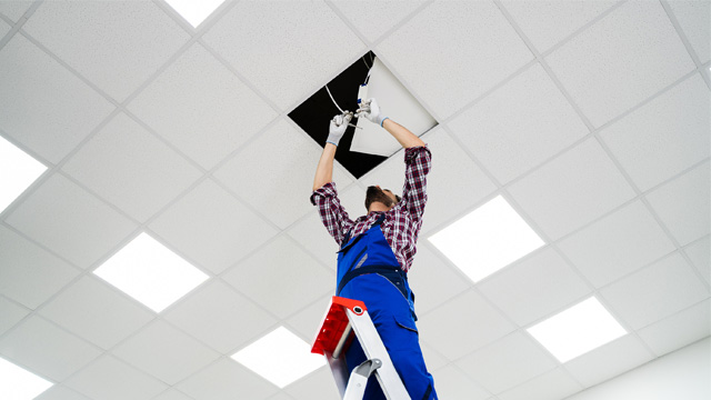 Electrician on stepladder installs lighting to the ceiling in office.
